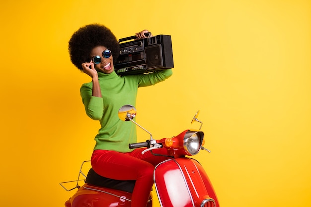 Ritratto fotografico di un elegante motociclista alla guida di una moto rossa che tiene in mano un boombox che tocca gli occhiali isolati su uno sfondo di colore giallo vivido