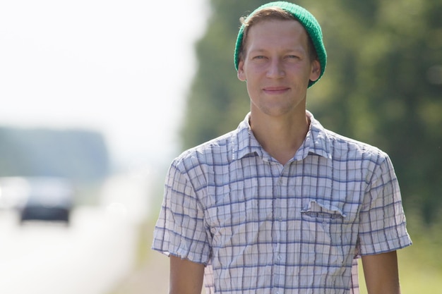 Ritratto fotografico di un giovane con una camicia a quadri e un cappello verde in piedi lungo la strada e sorride