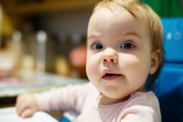 Ritratto fotografico di una bambina con le guance rosa si siede su una sedia per nutrirsi e mangia il porridge. Bambino seduto a casa