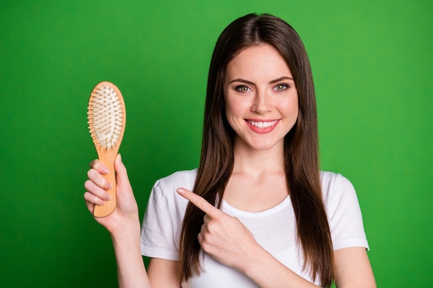 Ritratto fotografico di una ragazza che punta il dito contro la spazzola per capelli isolata su uno sfondo di colore verde vivido