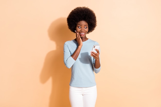 Ritratto fotografico di una ragazza dalla pelle scura impressionata che legge le informazioni dallo smartphone che tocca il viso con la mano isolata su uno sfondo di colore beige