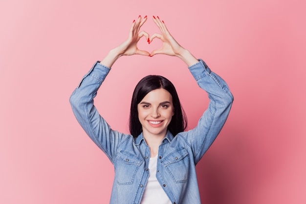 Ritratto fotografico di una ragazza romantica che mostra il cuore con le dita simbolo dell'amore il giorno di San Valentino isolato su sfondo di colore rosa pastello