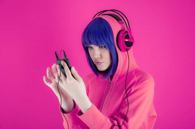 Ritratto funky bella giovane donna con i capelli blu in felpa con cappuccio su sfondo rosa brillante. La ragazza alternativa con le cuffie ascolta musica cool nel suo smartphone. Insolito concetto di moda giovanile. Immagine calda.