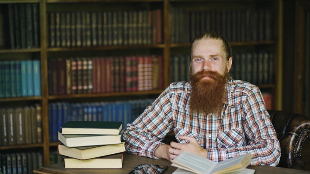 Ritratto giovane uomo barbuto sorridente felice in libreria e guardando nella fotocamera