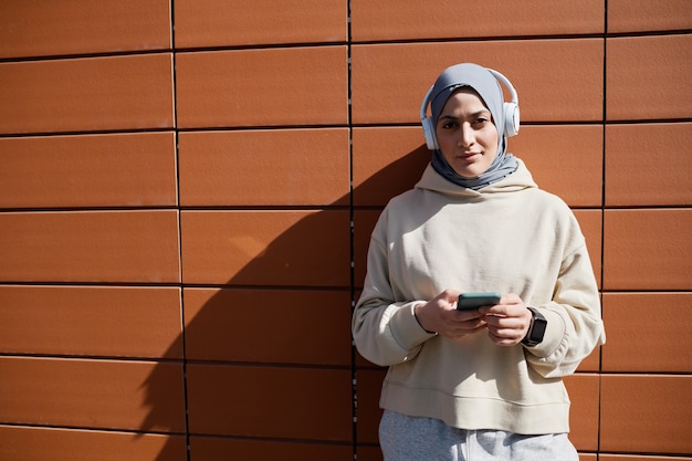 Ritratto grafico in vita di una donna mediorientale contemporanea che utilizza lo smartphone in città illuminata dalla luce del sole e indossa le cuffie, copia spazio