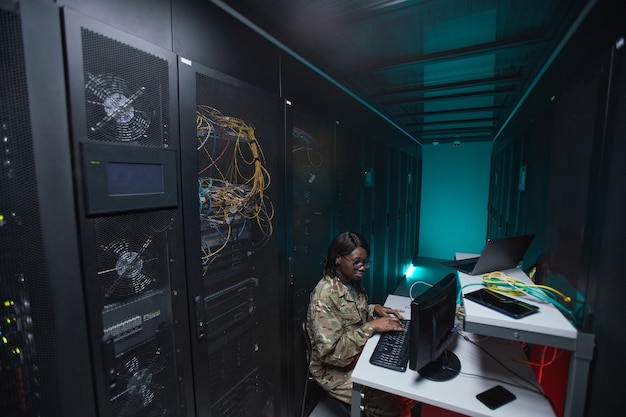 Ritratto grandangolare di giovane donna afro-americana che indossa l'uniforme militare che utilizza il computer durante la configurazione della rete nella sala server, spazio di copia