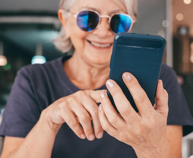 Ritratto in primo piano di una donna anziana sorridente con occhiali da sole seduta in un caffè usando uno smartphone