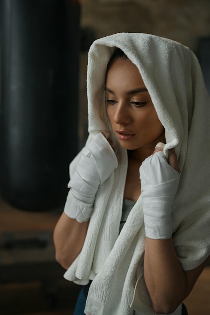 Ritratto in primo piano di una giovane pugile che indossa una benda da kickboxing sulle mani avvolte in un asciugamano e si sente stanca dopo l'allenamento.