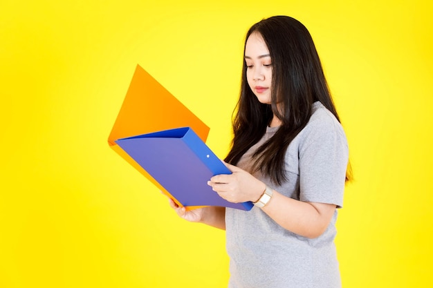 Ritratto in studio colpo di modello studentessa asiatica felice giovane paffuto grassoccio lungo capelli neri in abito casual in piedi sorridente che tiene le cartelle di file di documenti di scartoffie colorate su sfondo giallo.