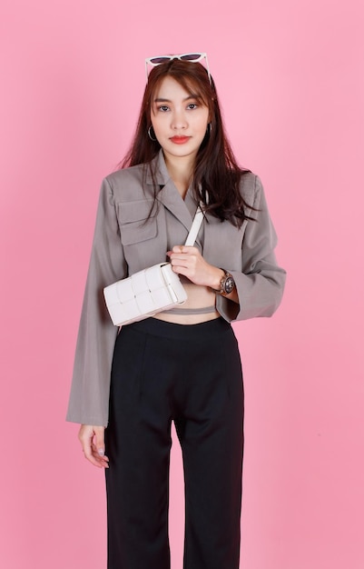 Ritratto in studio del modello teenager asiatico alla moda hipster femminile in casual crop top street indossa occhiali da sole giacca che trasportano borsa in pelle borsa in piedi guarda la fotocamera su sfondo rosa.