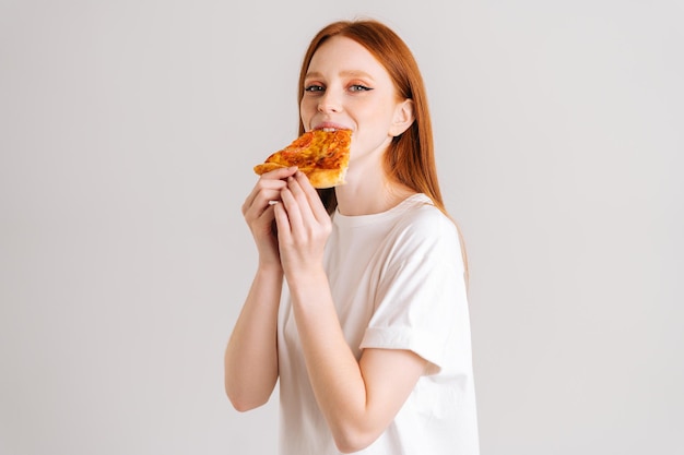 Ritratto in studio di allegra giovane donna felice con appetito per mangiare una deliziosa pizza guardando la fotocamera in piedi su sfondo bianco isolato. Femmina graziosa della testarossa che mangia pasto saporito.