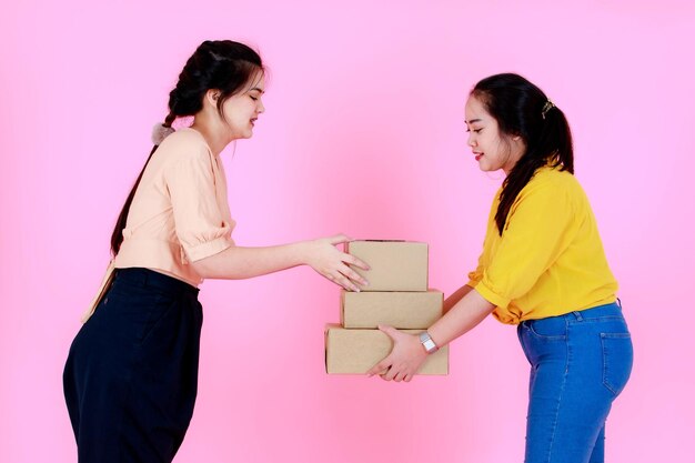 Ritratto in studio di due asiatiche paffute e sottili bretelle denti coda di cavallo capelli partner d'affari femminile in abbigliamento casual in piedi sorridente che tiene la spedizione del pacchetto di consegna insieme su sfondo rosa.