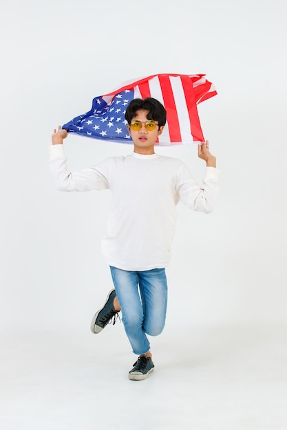 Ritratto in studio di giovane asiatico LGBT gay bisessuale omosessuale maschio modello alla moda in abito casual e occhiali da sole alla moda in piedi tenendo sventola bandiera nazionale USA volante su sfondo bianco