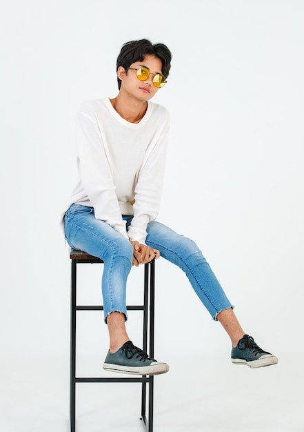 Ritratto in studio di giovane asiatico LGBT gay felice bisessuale omosessuale maschio modello alla moda in abbigliamento casual moda occhiali da sole in piedi appoggiato alla sedia alta sorridente in posa su sfondo bianco