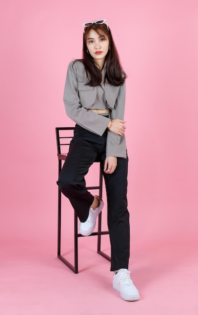 Ritratto in studio di giovane asiatico urbano alla moda femminile hipster adolescente modello di moda in casual crop top street indossa giacca con occhiali da sole sulla testa seduta guardare la fotocamera su sfondo rosa
