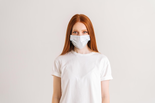 Ritratto in studio di giovane donna sicura di sé che indossa una maglietta casual in maschera protettiva guardando la fotocamera in piedi su sfondo bianco isolato epidemia di influenza allergia alla polvere protezione contro i virus