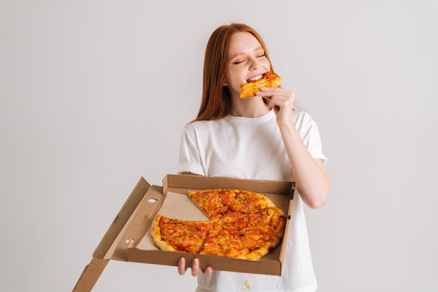 Ritratto in studio di giovane donna soddisfatta con gli occhi chiusi con appetito per mangiare una deliziosa pizza tenendo la scatola in mano in piedi su sfondo bianco isolato Bella femmina rossa che mangia un pasto gustoso