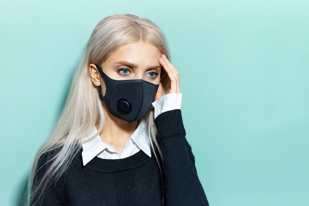 Ritratto in studio di giovane ragazza bionda, avendo mal di testa, indossando maschera respiratoria di colore nero, contro il virus. Sfondo di colore ciano, aqua menthe.
