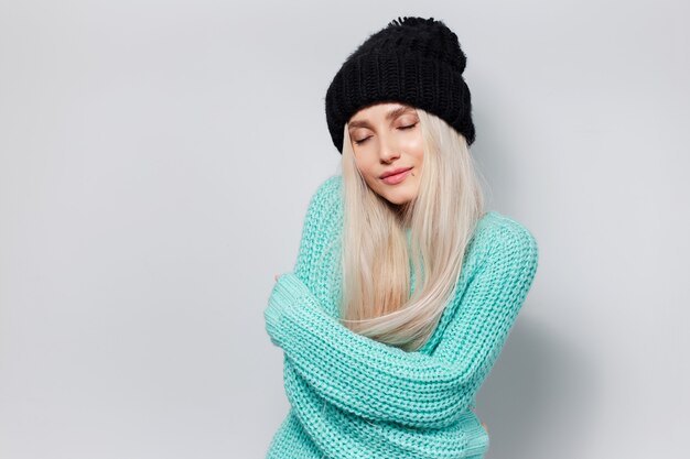 Ritratto in studio di giovane ragazza bionda carina che si abbraccia su sfondo bianco. Indossa un cappello nero e un maglione ciano.