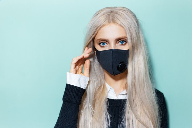 Ritratto in studio di giovane ragazza bionda che indossa una maschera respiratoria nera contro il coronavirus sul colore dell'acqua.