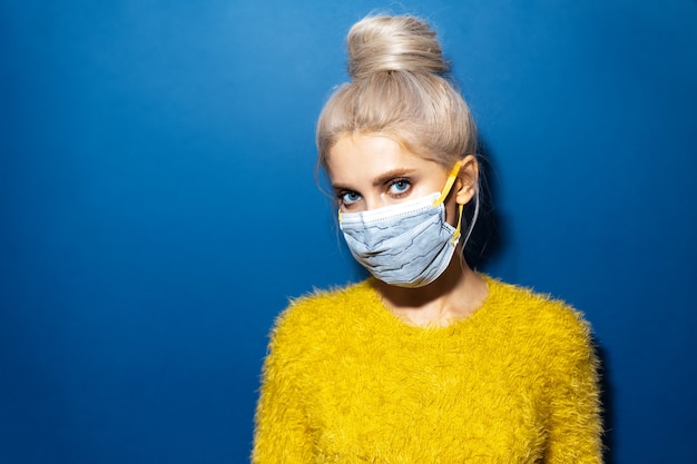 Ritratto in studio di giovane ragazza bionda con chignon per capelli che indossa maschera di sicurezza respiratoria medica sul viso contro il coronavirus sul blu