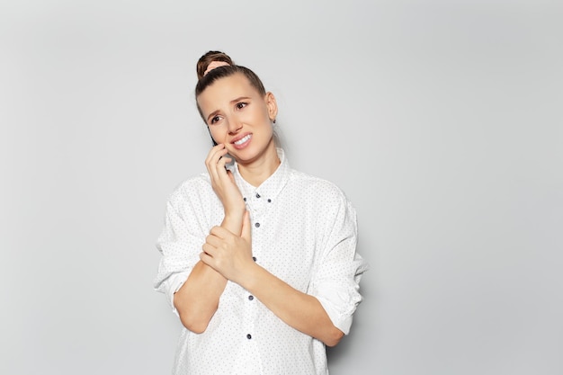 Ritratto in studio di giovane ragazza felice deludente con chignon per capelli che parla su smartphone su sfondo grigio guardando in su indossando una camicia bianca