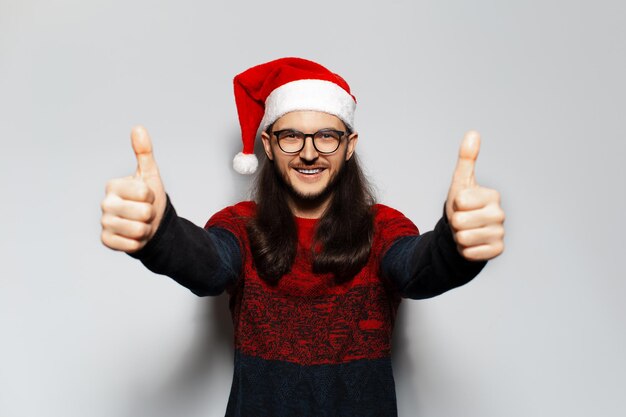 Ritratto in studio di giovane uomo felice con capelli lunghi e occhiali da vista che mostra i pollici in su indossa maglione rosso di Natale e cappello di Babbo Natale su sfondo bianco