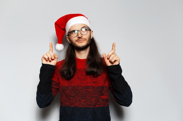 Ritratto in studio di giovane uomo sorridente bello con i capelli lunghi che puntano il dito indice verso l'alto indossa maglione rosso di Natale e cappello di Babbo Natale su sfondo bianco