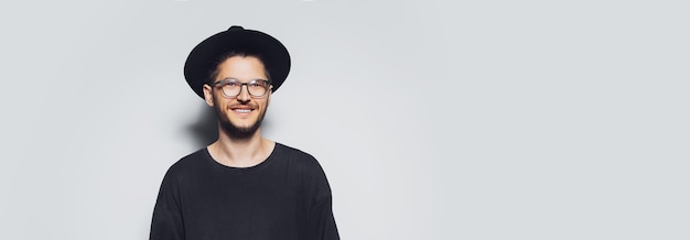 Ritratto in studio di giovane uomo sorridente che indossa occhiali da vista e cappello nero su sfondo grigio con spazio per la copia Vista panoramica del banner