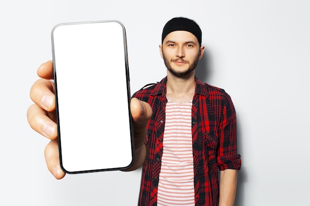 Ritratto in studio di giovane uomo sorridente che tiene in mano un grande smartphone con schermo vuoto che mostra vicino alla fotocamera un dispositivo con mockup su sfondo bianco che indossa una maglietta rossa