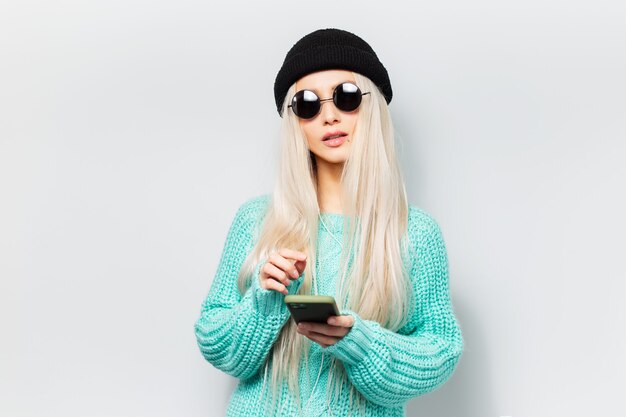 Ritratto in studio di ragazza bionda giovane hipster utilizzando smartphone su sfondo bianco. Indossare occhiali da sole rotondi e cappello a cuffia nero.