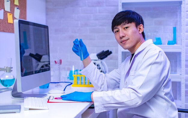 Ritratto in studio di scienziato maschio professionista asiatico in guanti di gomma da laboratorio bianco seduto sorridente sguardo alla fotocamera che mostra la provetta del campione nelle mani nella sala di lavoro del laboratorio dell'ospedale.