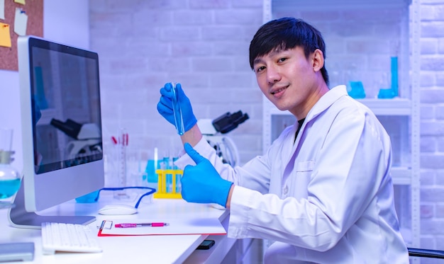 Ritratto in studio di scienziato maschio professionista asiatico in guanti di gomma da laboratorio bianco seduto sorridente sguardo alla fotocamera che mostra la provetta del campione nelle mani nella sala di lavoro del laboratorio dell'ospedale.