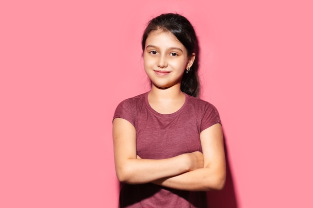 Ritratto in studio di sorridente ragazza bambino con le braccia incrociate su sfondo di colore rosa pastello.