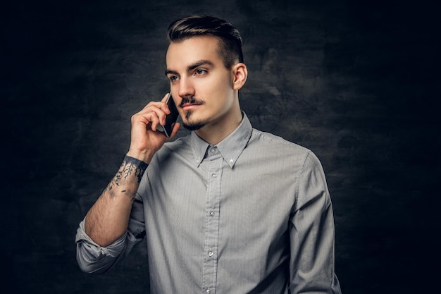 Ritratto in studio di un bel maschio hipster barbuto con un tatuaggio sul braccio parla su uno smartphone.