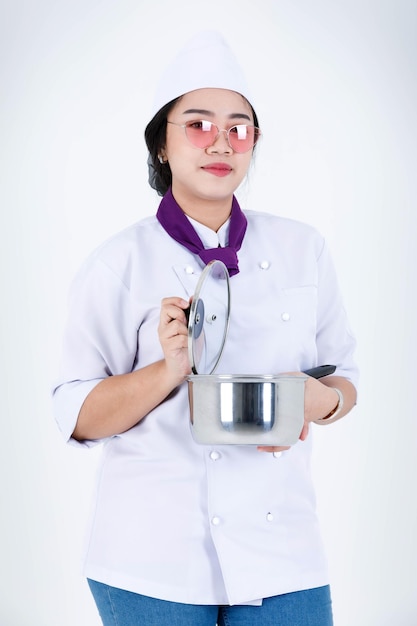 Ritratto in studio di un ristorante asiatico professionale che cucina chef esecutivo femminile in uniforme da cuoco e sciarpa in piedi sorridente guarda la telecamera che tiene pentola in acciaio inossidabile e coperchio su sfondo bianco.