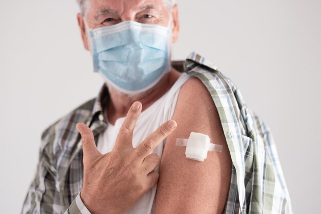 Ritratto in studio di un vecchio paziente caucasico anziano che indossa una maschera facciale che mostra con le dita numero tre come terze dosi di vaccino contro il coronavirus covid-19, guardando la fotocamera