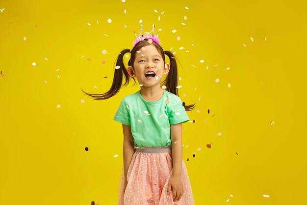 Ritratto in studio di una bambina gioiosa che celebra allegramente il suo compleanno con confetti su una parete gialla