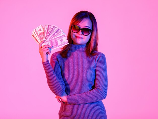 Ritratto in studio di una giovane asiatica ricca e ricca modello femminile cool in maglione a collo alto grigio e occhiali da sole con lenti nere che tengono una pila di banconote da cento dollari piena su sfondo rosa chiaro.