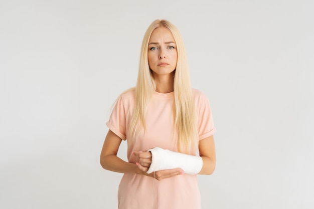 Ritratto in studio di una giovane donna bionda ferita infelice con il braccio rotto avvolto in una benda di gesso che guarda l'obbiettivo in piedi su sfondo bianco isolato