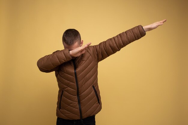 Ritratto in studio. Giovane 20s uomo in una giacca marrone, su sfondo giallo, facendo una pennellata di danza hip-hop mani si muove gesto segno giovanile nascondere il viso