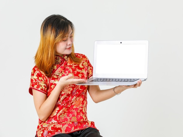 Ritratto in studio girato modello femminile asiatico millenario in camicia rossa cinese tradizionale cheongsam qipao seduto che tiene mostrando la presentazione del prodotto del computer portatile schermo vuoto su sfondo bianco.