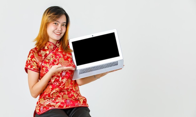 Ritratto in studio girato modello femminile asiatico millenario in camicia rossa cinese tradizionale cheongsam qipao seduto tenendo mostrando che presenta prodotto di computer portatile schermo vuoto nero su sfondo bianco.