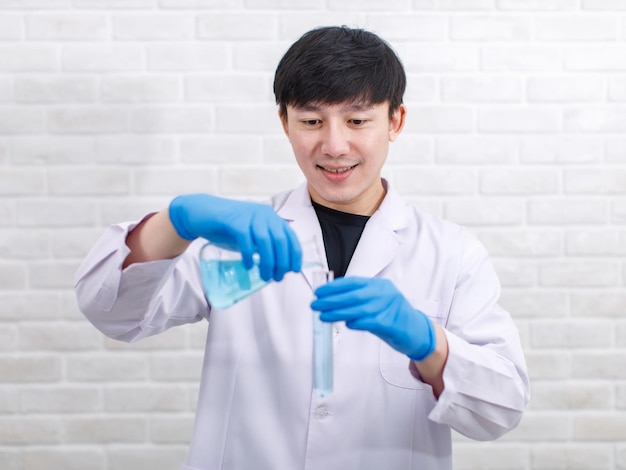 Ritratto in studio Scienziato maschio professionista asiatico in guanti di gomma da laboratorio bianco in piedi, sorridente, guardare la fotocamera tenendo la vetreria vuota vuota del matraccio di Erlenmeyer nelle mani su sfondo muro di mattoni.