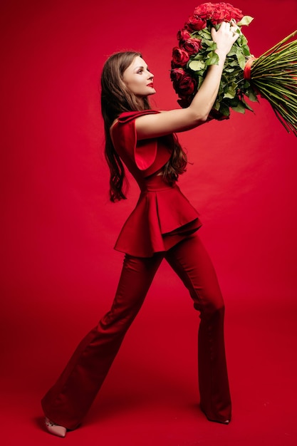 Ritratto in studio vista laterale di una splendida giovane bruna con i capelli lunghi che indossa un abito rosso alla moda Sta sorridendo al grande mazzo di rose rosse nelle sue mani Colore rosso totale Isolare su sfondo rosso