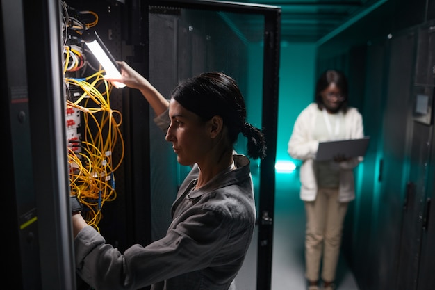 Ritratto in vista laterale dell'ingegnere di rete femminile che collega i cavi nell'armadio del server mentre si lavora con il supercomputer nel data center, spazio di copia