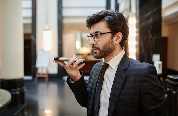 Ritratto in vista laterale di un uomo d'affari di successo che registra un messaggio vocale sullo smartphone mentre si trova nella hall dell'hotel, copia spazio