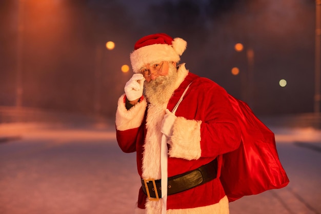 Ritratto in vita del tradizionale Babbo Natale in piedi nella strada della città di notte e guardando la fotocamera