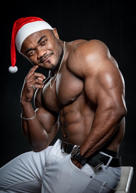 Ritratto in vita dell'uomo bodybuilder afroamericano con torso muscoloso senza maglietta. Maschio che indossa il cappello di Natale isolato su sfondo nero