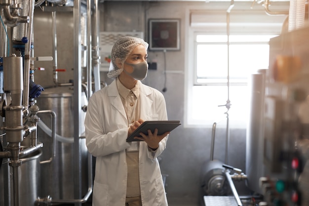 Ritratto in vita di giovane donna che indossa maschera e tiene in mano un tablet digitale durante l'ispezione del controllo di qualità in un'officina illuminata dal sole presso una fabbrica di alimenti, copia spazio